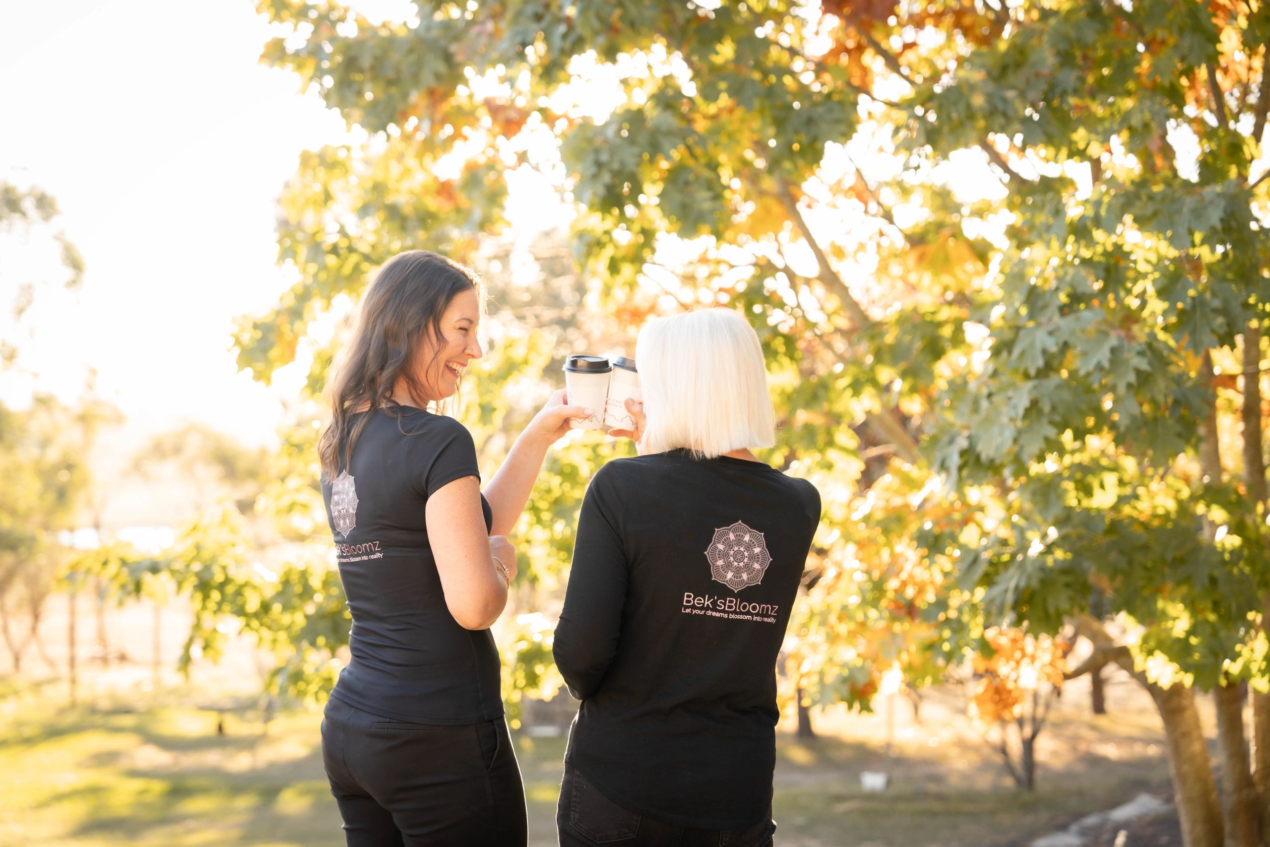 Two women clinking coffee cups outdoors on a sunny day.