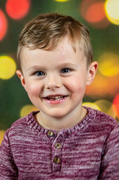 Little boy wearing a read and white sweater,  sitting in front of colorful lights smiling. 