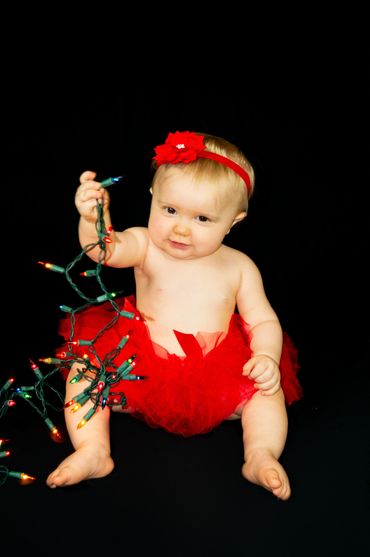 Little baby girl on a black backdrop. She is wearing a red tutu and a red flower headband holding a 