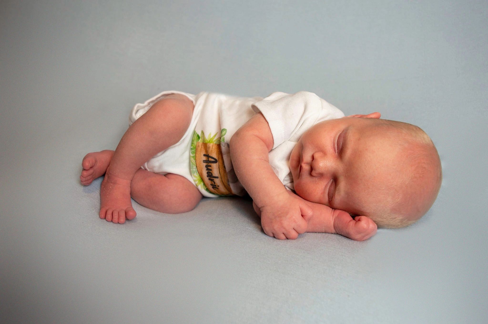 Infant baby boy wearing white one piece, curled up and sleeping.