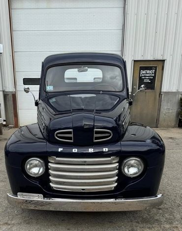Front view of a classic black Ford truck parked outside an automotive shop.