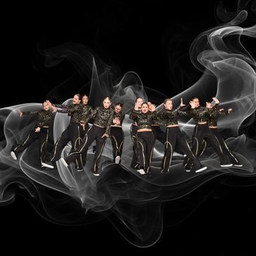 A group of dancers in black and gold outfits performing against a smoky black background.