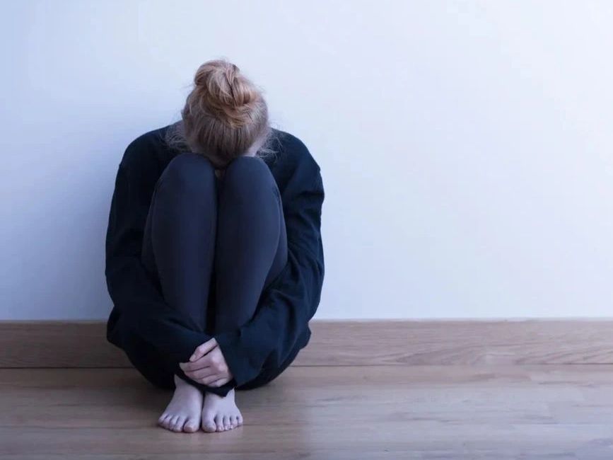 Person sitting on floor hugging knees, head down.
