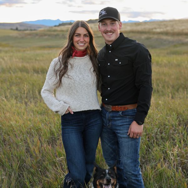 Smiling couple with a dog in a grassy field at sunset.
