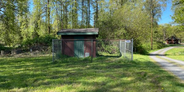 Small fenced utility shed in a green forest clearing under blue sky.