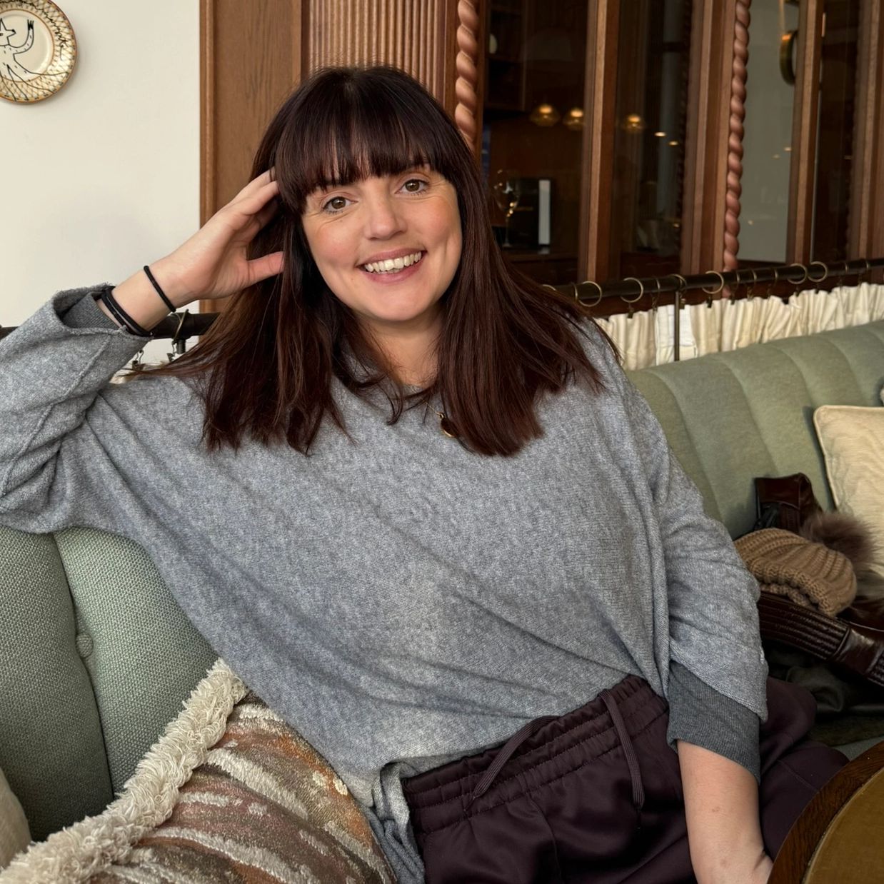A woman in a gray sweater smiling while sitting on a sofa indoors.