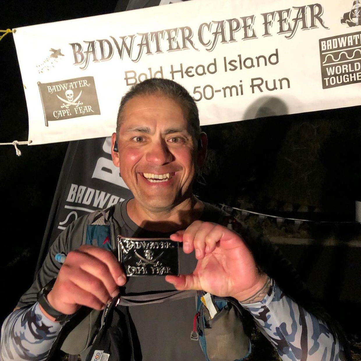 Smiling man proudly holds a Badwater Cape Fear award at night.