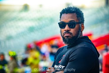Man with sunglasses and a beard in a stadium.