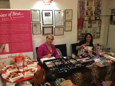 Two women at a market stall displaying various accessories and books.