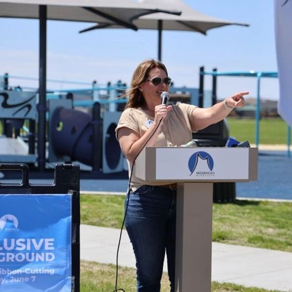 Woman speaking at an inclusive playground ribbon-cutting event outdoors.