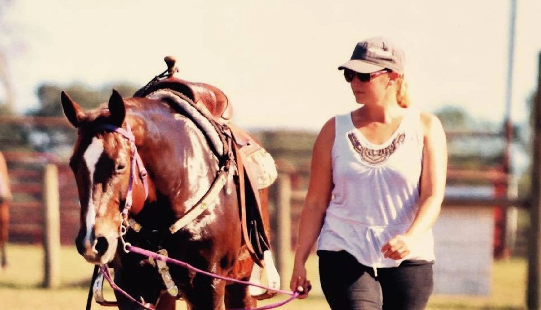 Woman walking a saddled horse outdoors on a sunny day.