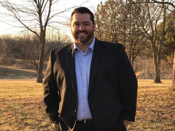 A smiling man in a suit standing outdoors in a sunlit park.
