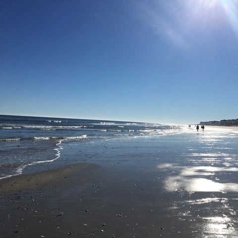 Sunny beach with gentle waves and a few people walking along the shore.