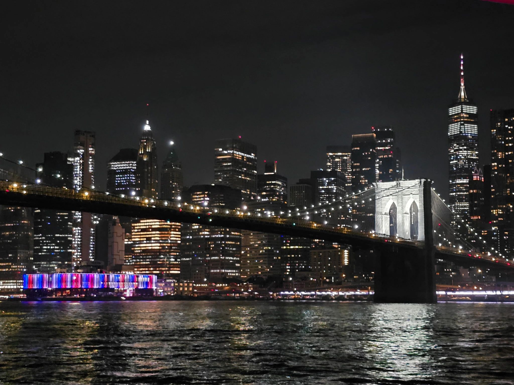 NYC skyline at night from the water featuring Brooklyn Bridge and One World Trade Center