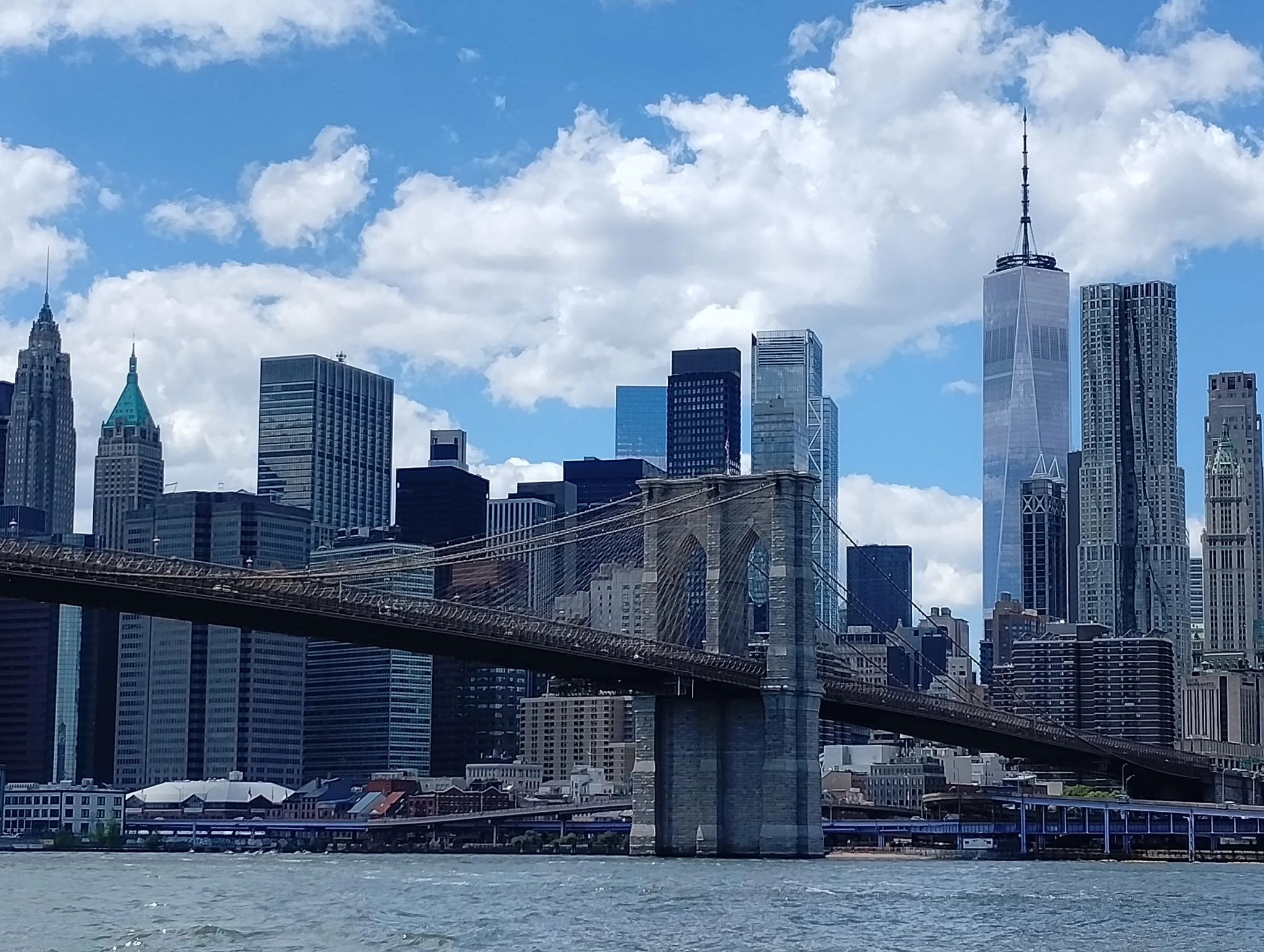 Brooklyn Bridge with Manhattan skyline under a blue, partly cloudy sky.