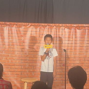 Young boy performing stand-up comedy on stage with microphone.
