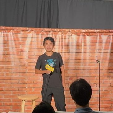 Young comedian performing stand-up on stage with mic and stool.