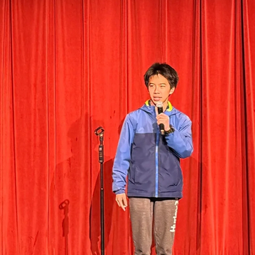 Young man speaking on stage with a microphone in front of red curtains.