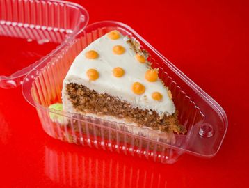 A slice of carrot cake with white frosting and orange dots in a plastic container.