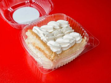 A slice of cake with white frosting in a plastic container on a red surface.