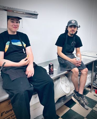 Two smiling employees sitting on a steel prep table while taking a break.  
