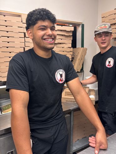 Two smiling employees stand before multiple folded pizza boxes.  
