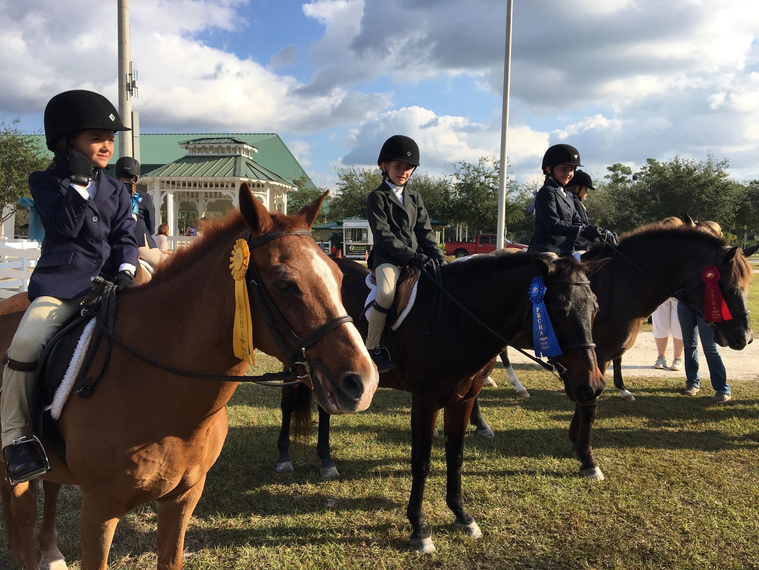 Young riders with ribbon-winning horses at an outdoor equestrian event.