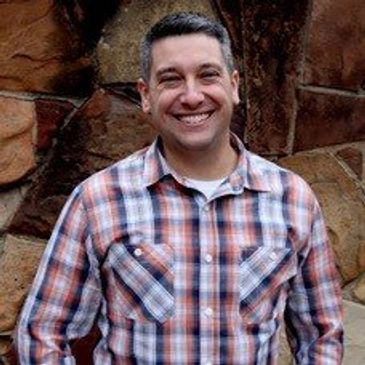 Smiling man in a plaid shirt standing against a stone wall.
