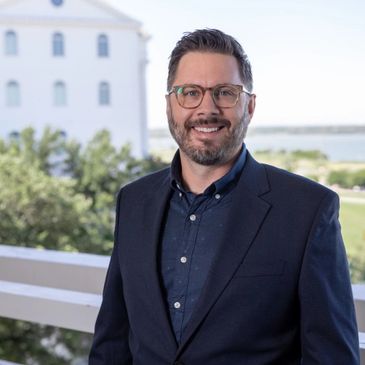 Smiling man in glasses and blazer standing outdoors with a scenic background.
