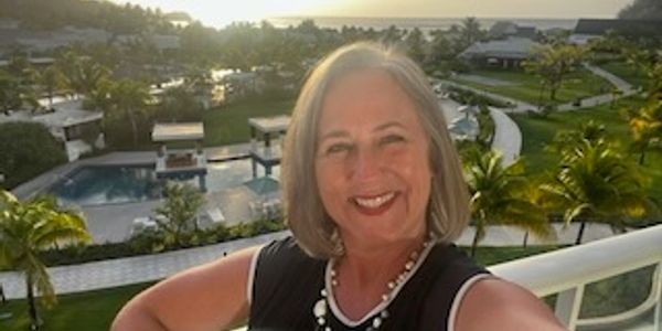 Smiling woman taking a selfie on a balcony with a tropical resort sunset in the background.