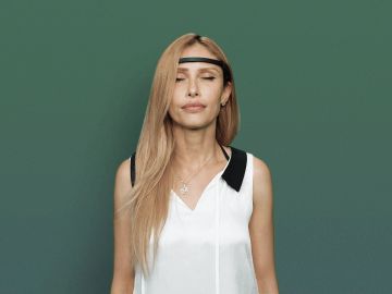 Woman with long blonde hair wearing a white top and black headband against a green background.