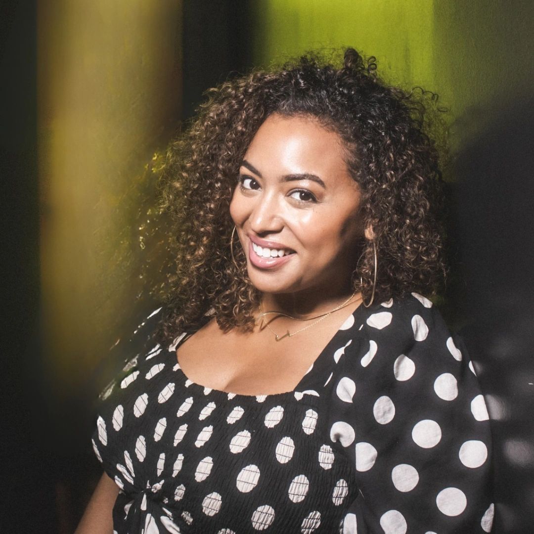 Smiling woman with curly hair wearing a black polka dot dress.