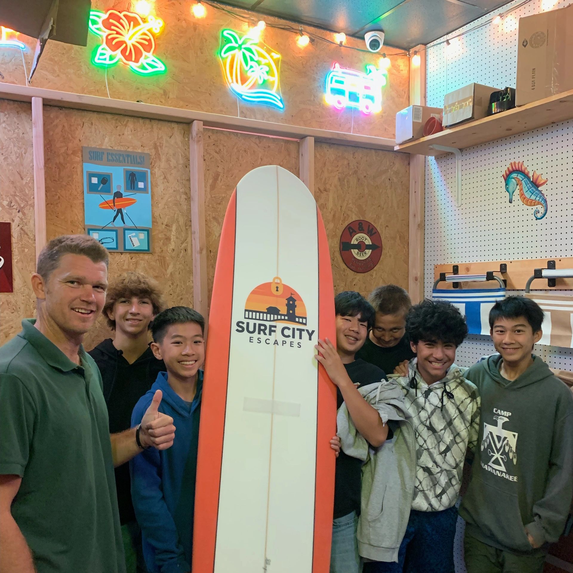 Group of boys and a man posing with a surfboard indoors, colorful neon signs decorate the wall.
