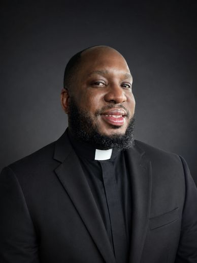 Portrait of a smiling Black clergyman in a black suit with a white clerical collar.