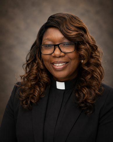 A smiling woman wearing glasses and clerical collar with curly hair.