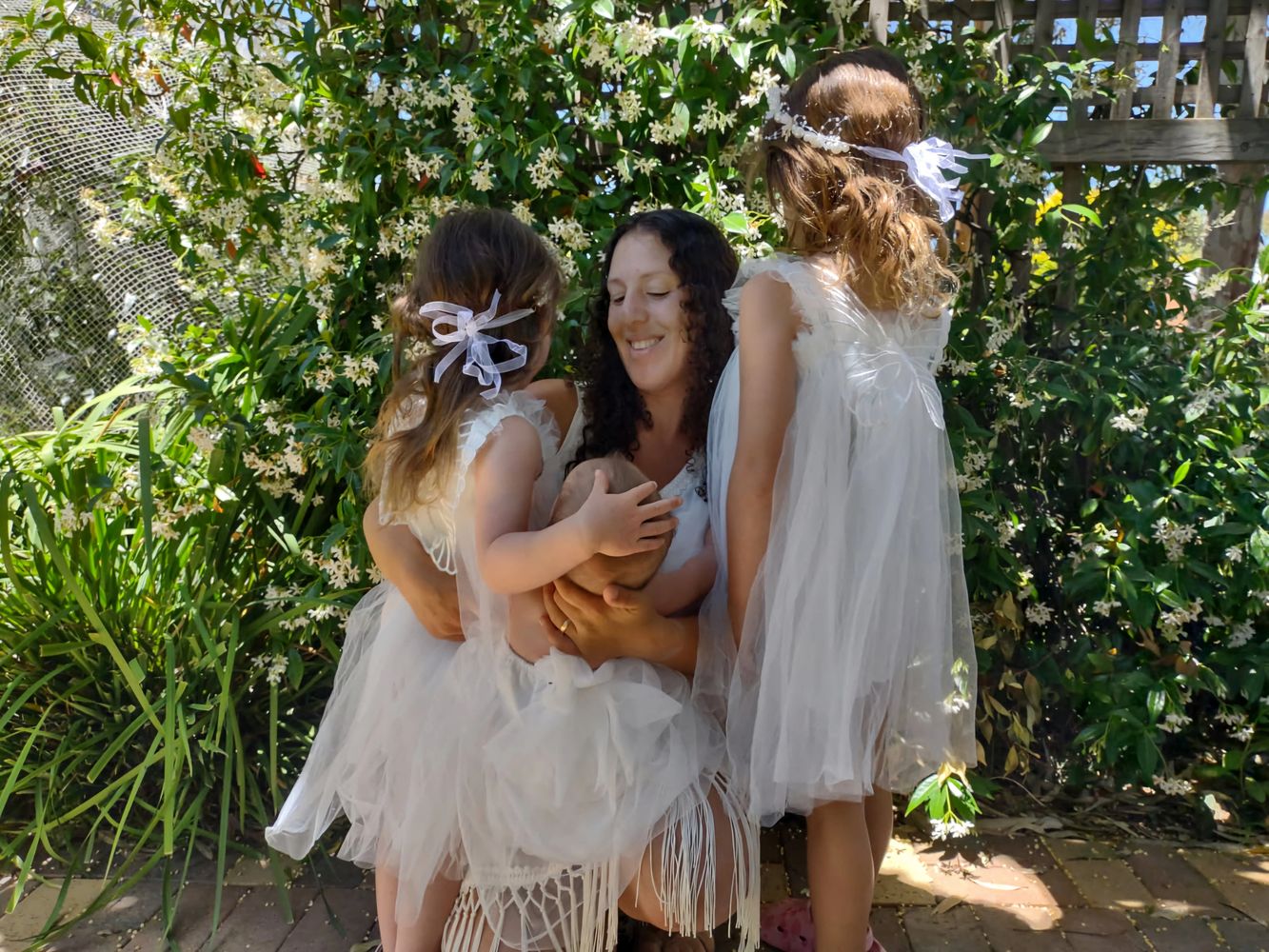 A joyful mother embraces her children dressed in white dresses in a garden.