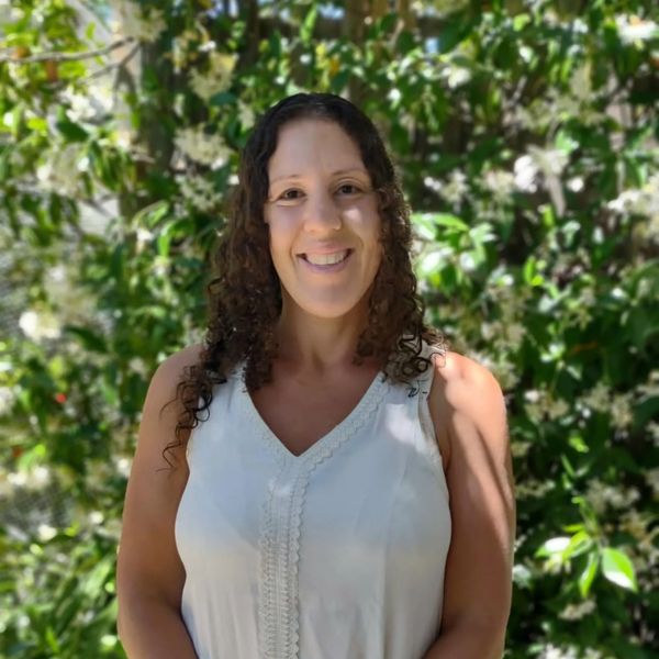 A smiling woman in a white sleeveless top with curly hair stands outdoors in front of a green leafy background.