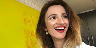 A smiling woman with ombre hair holds a marker near a yellow board with writing.