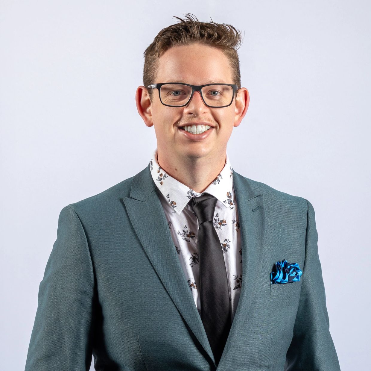 Man in a teal suit with floral shirt and glasses smiles at the camera.