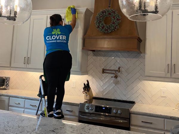 A cleaner in a blue shirt wipes a kitchen cabinet using a step stool.