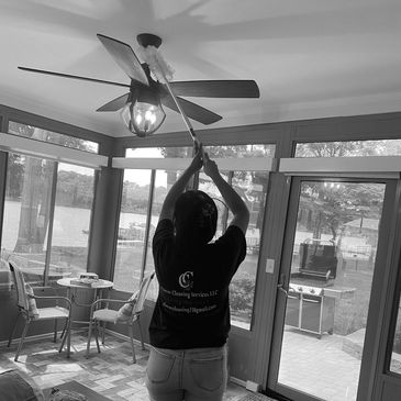 Person dusting a ceiling fan in a sunroom with large windows.