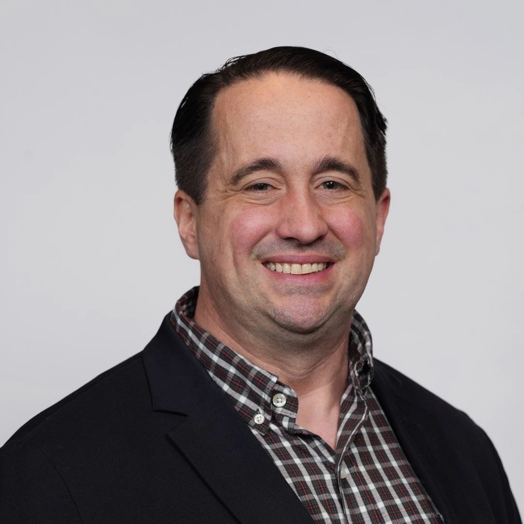Smiling man in a checked shirt and black blazer against a plain background.