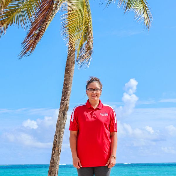 A smiling woman in a red shirt stands by a palm tree on a sunny beach.