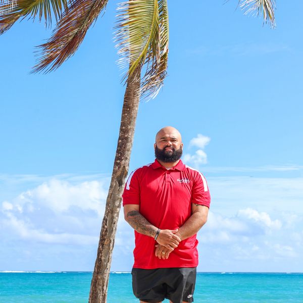 Man in red shirt standing by a palm tree on a sunny beach.