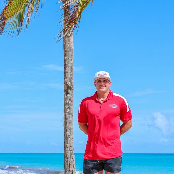Man in red shirt stands by a palm tree on a tropical beach.
