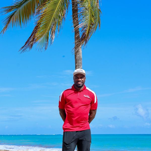 Man in red shirt standing on the beach under a palm tree with blue sky.
