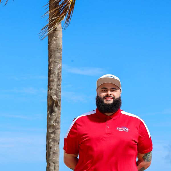 Man with a beard in a red shirt standing by a palm tree under a clear blue sky.