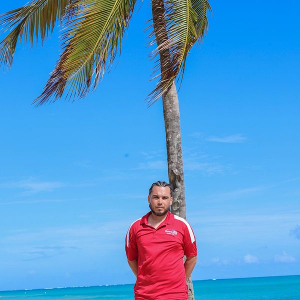Man in red shirt standing by a palm tree on a sunny beach.