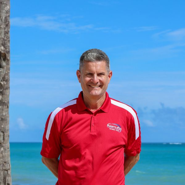 Man smiling in a red Puerto Rico National Team shirt by the ocean.