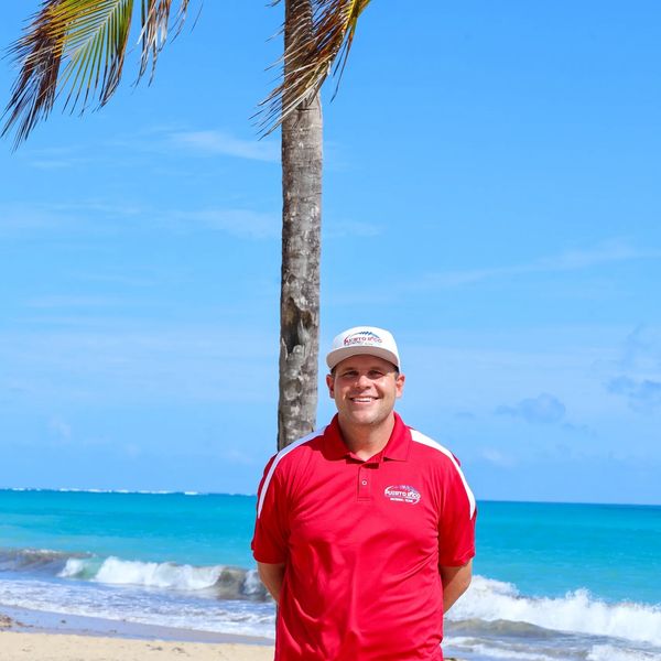 Man in red shirt smiling near a palm tree on a beach.